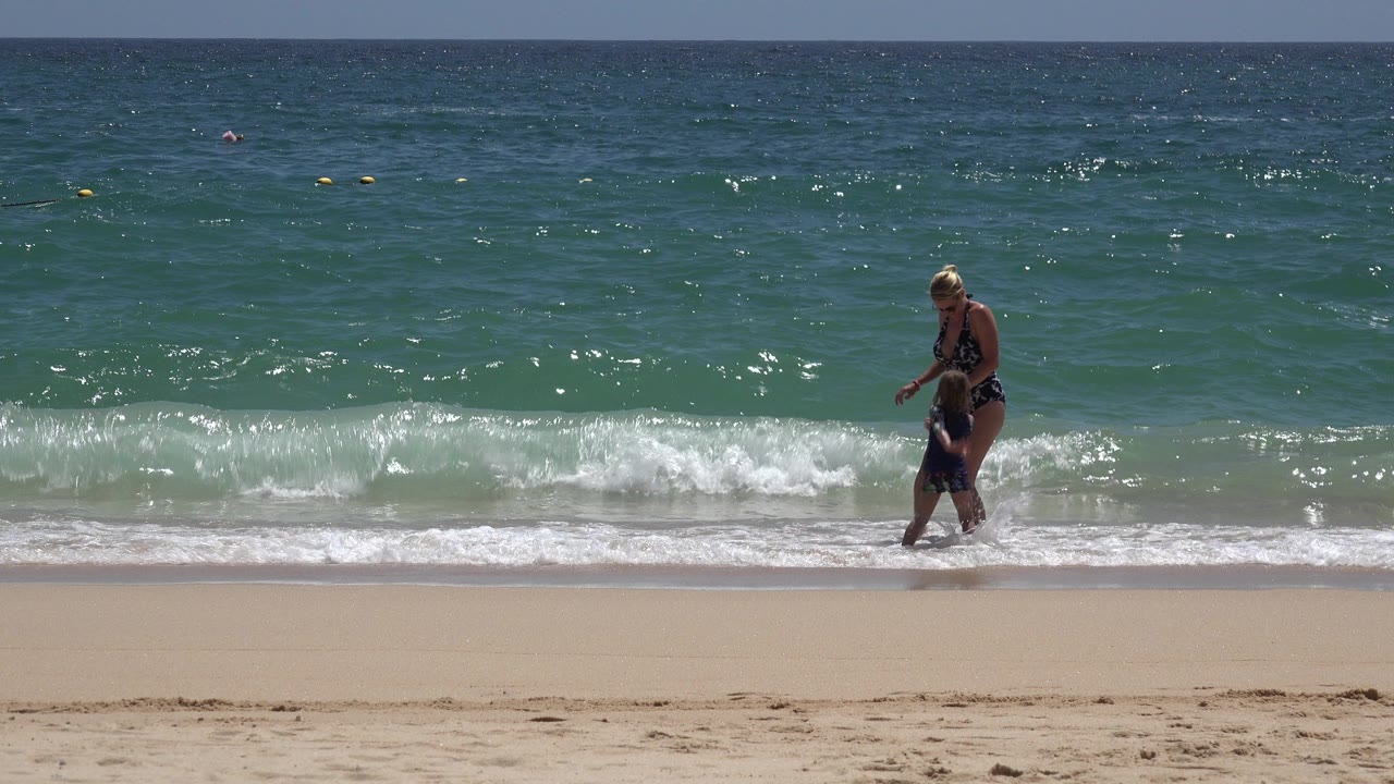 The adult woman and child remain near the shore in Cabo San Lucas, Mexico, while a low white wave spreads across the waterline behind them.