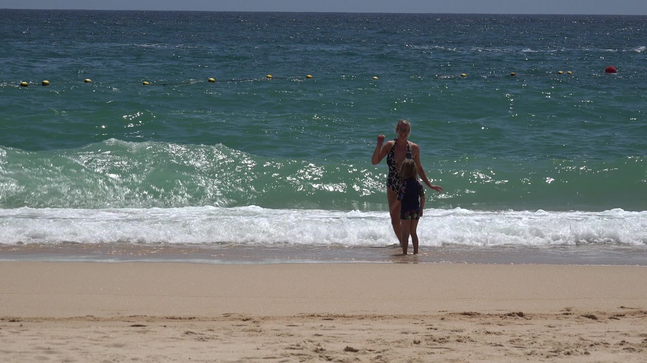 At 0:01 in Cabo San Lucas, Mexico, the woman is centered near the waterline while white foam spreads along the shore behind her.