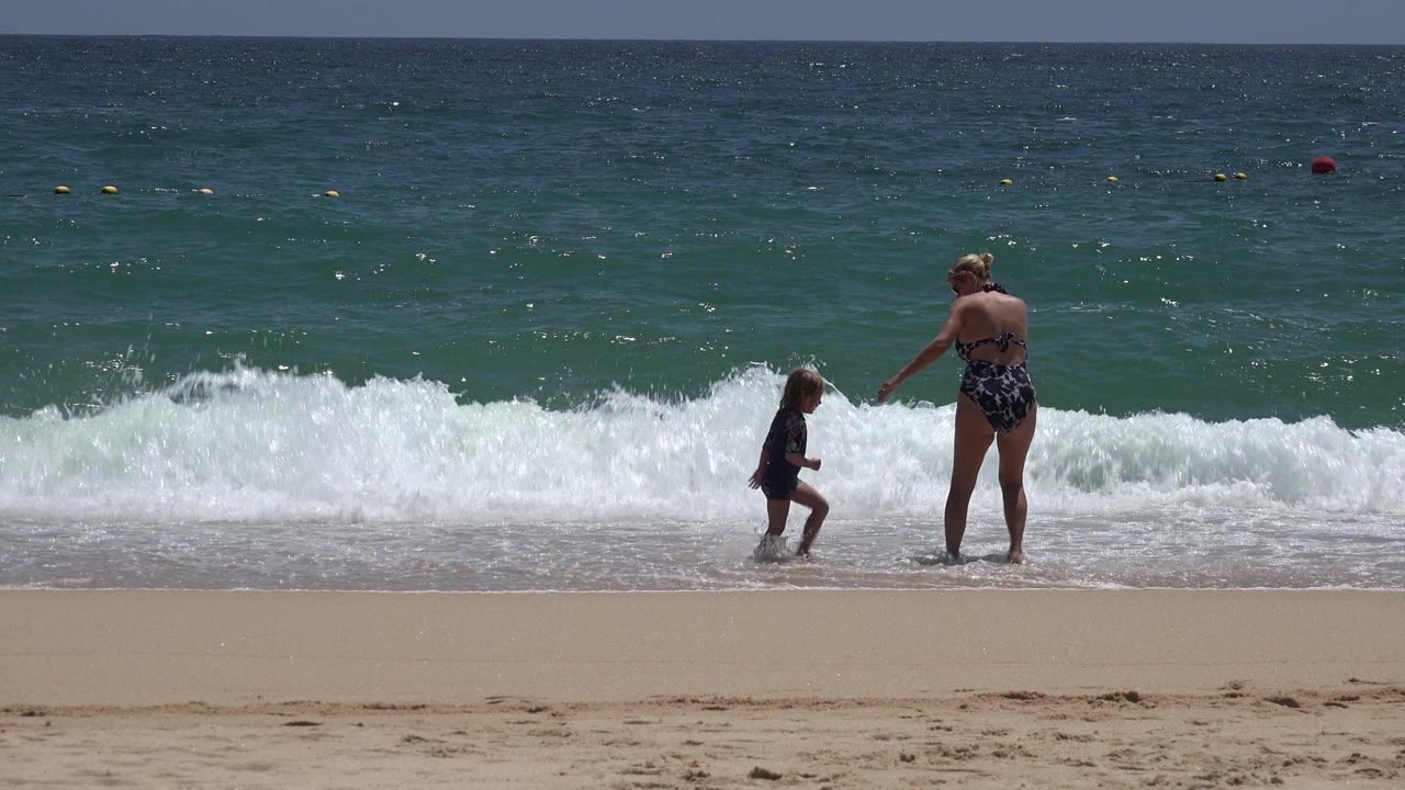 Woman and child standing near breaking surf on a sandy beach.