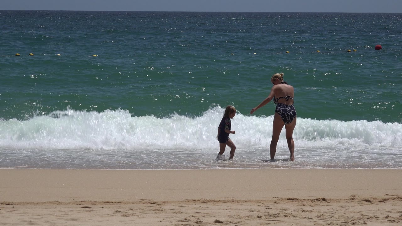The child steps along the wet sand while the woman reaches an arm toward them in Cabo San Lucas, Mexico, with a broad white wave cresting behind them.