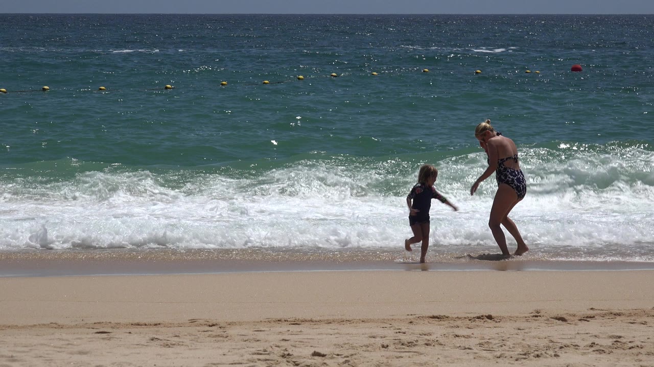In Cabo San Lucas, the child moves toward the woman while a low wave spreads white foam across the shoreline behind them.