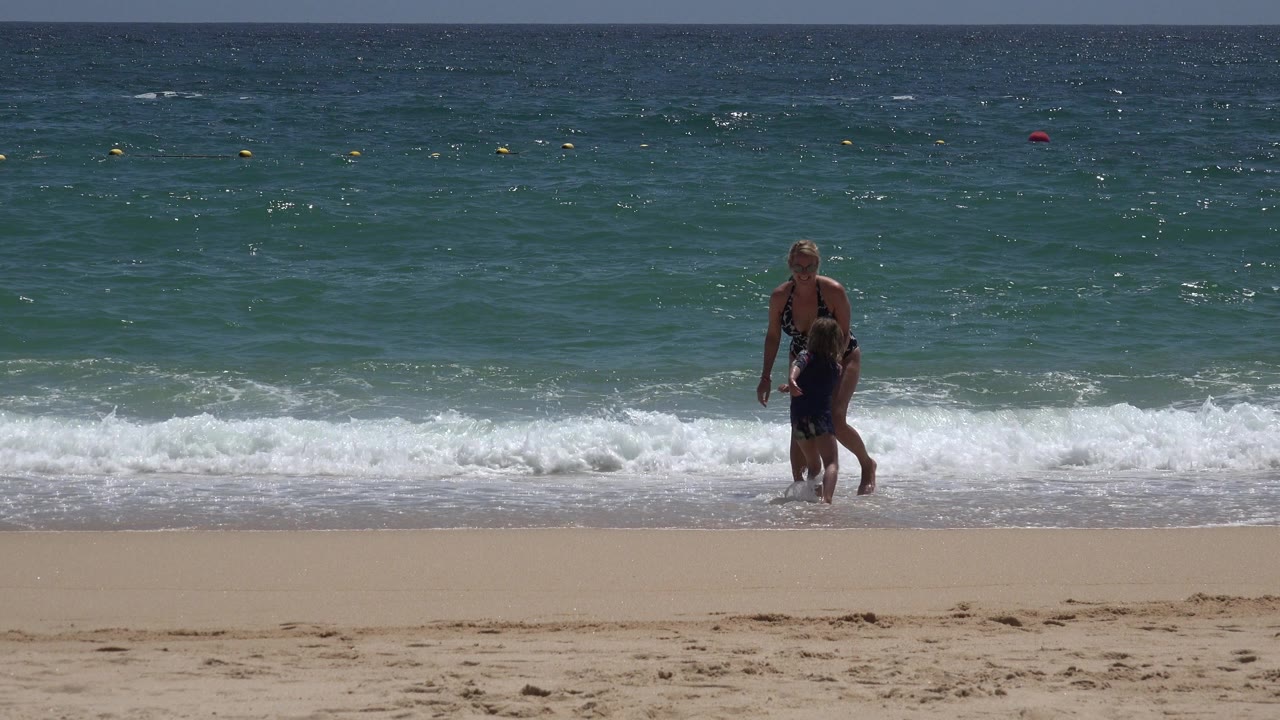 A woman and young child stand near the breaking waves on a sandy Cabo San Lucas beach, with green-blue ocean and buoy markers in the background.