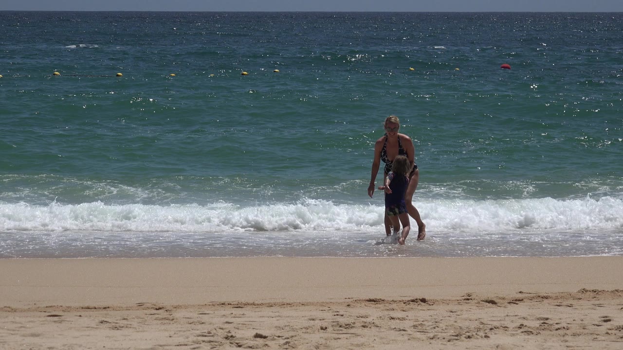 The woman and child stay close together at the Cabo San Lucas waterline while a low white wave spreads across the shore behind their legs.