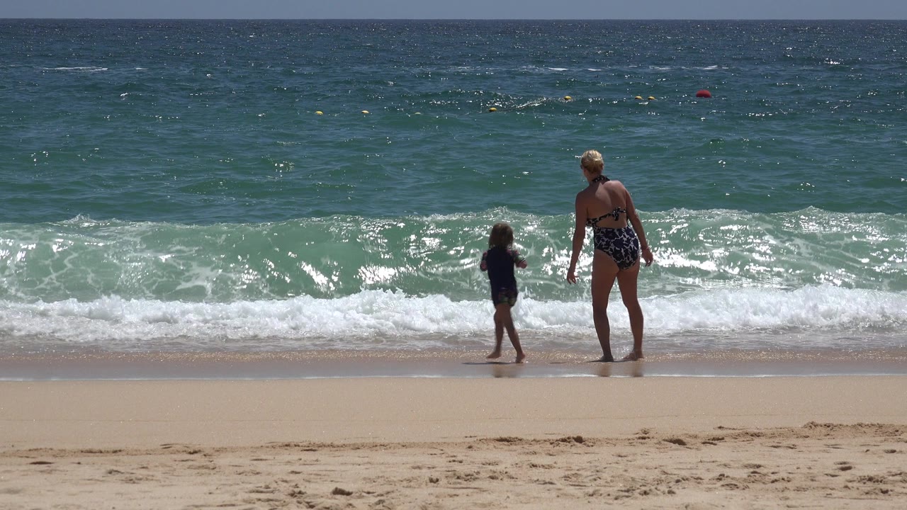 Child and adult standing at the shoreline as a wave approaches.