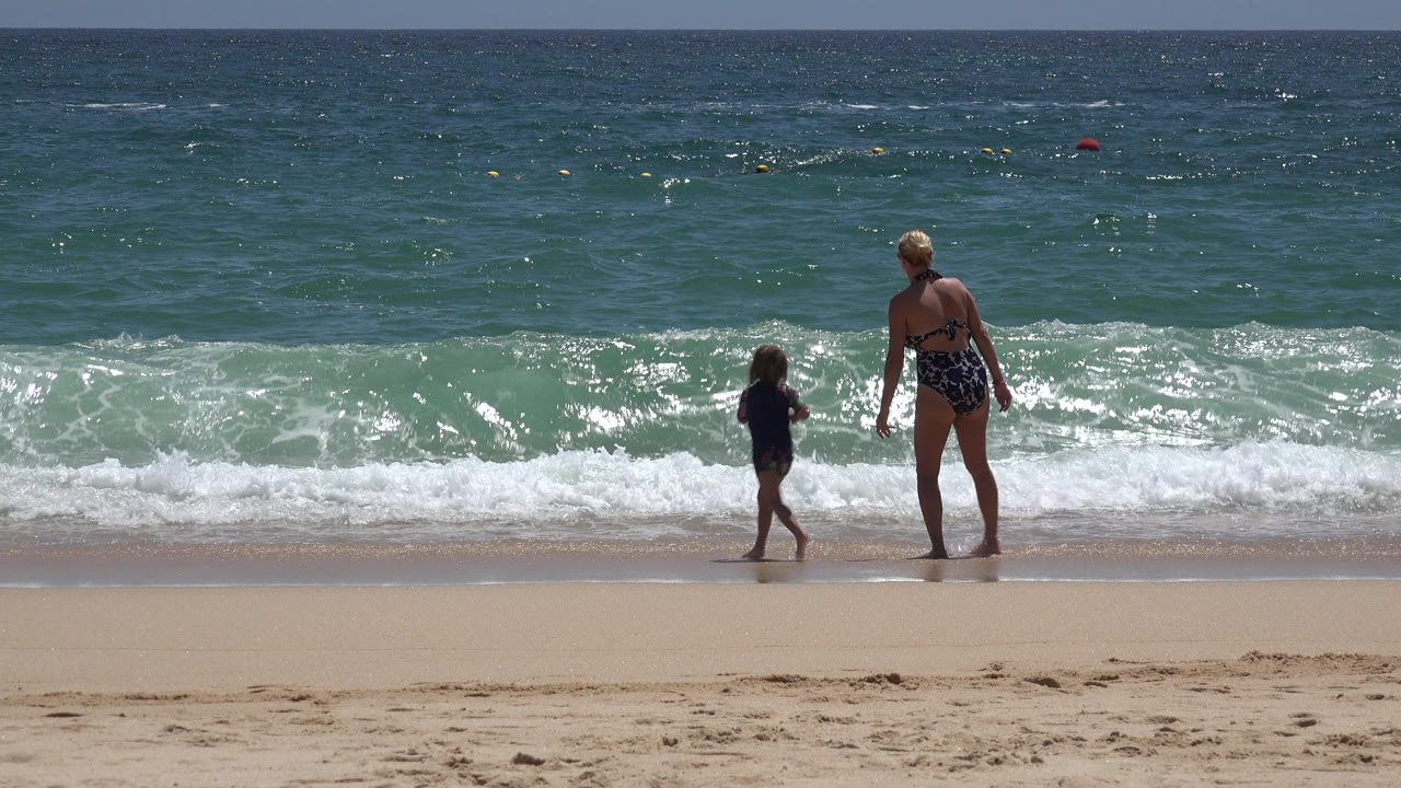In Cabo San Lucas, Mexico, the child steps along the wet shoreline while the adult stands nearby and white surf spreads across the sand.