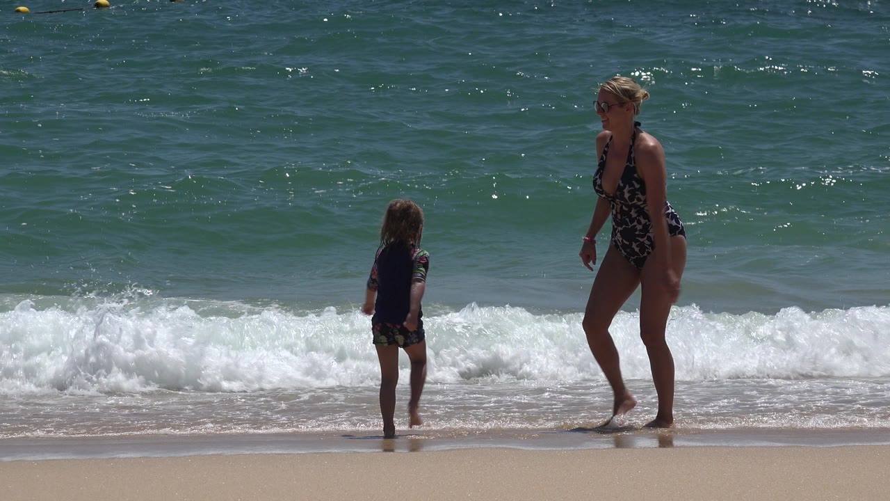 The child faces the incoming water while the mother pauses a few steps away on the Cabo San Lucas shoreline, with sun highlights on the ocean surface.
