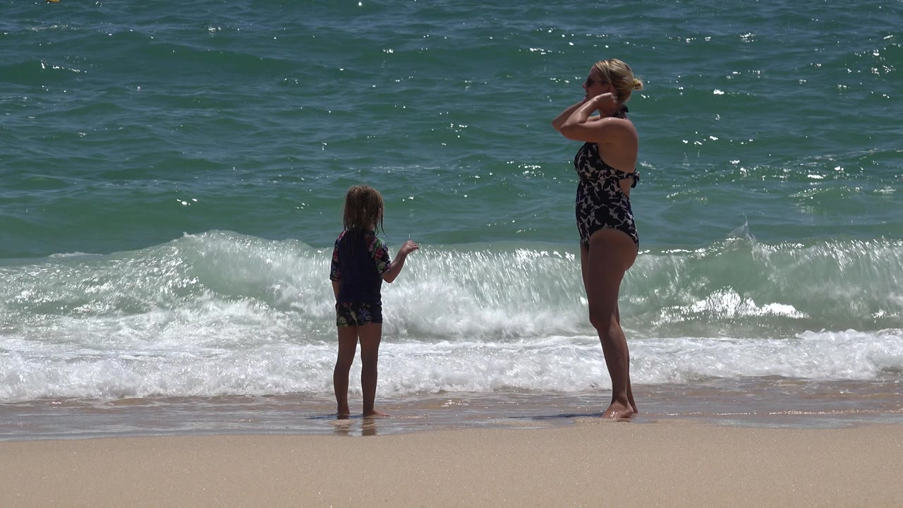 A woman in black swimwear and a young girl stand on the wet sand at the surf line in Cabo San Lucas, Mexico, with small waves breaking behind them.