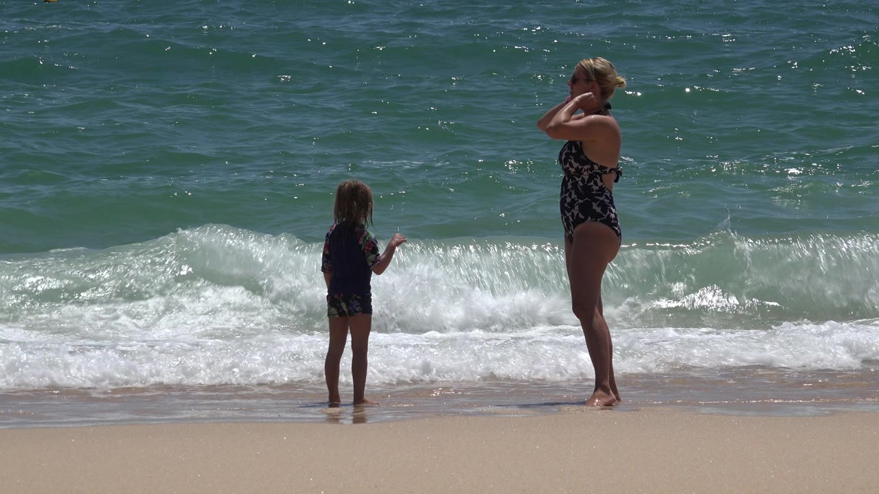 The woman adjusts her hair while the young girl faces the water in Cabo San Lucas, Mexico, with foamy surf spreading across the sand.