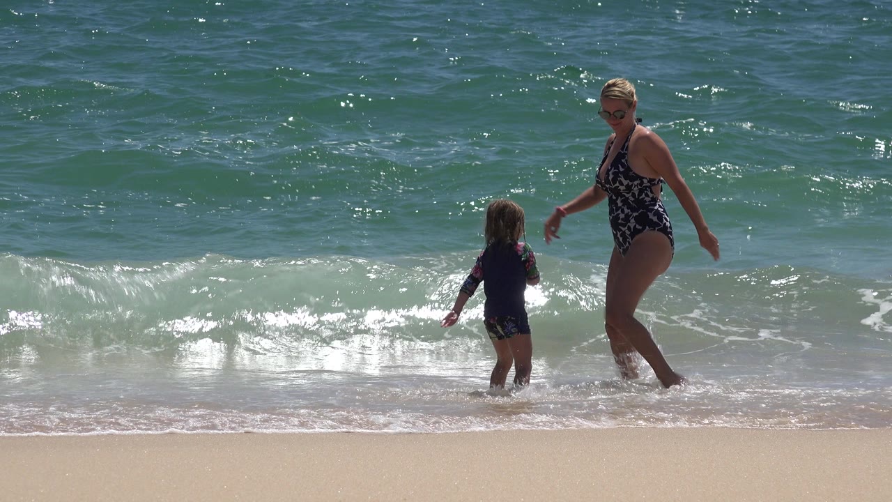 Woman and child standing in shallow surf at a sandy beach.