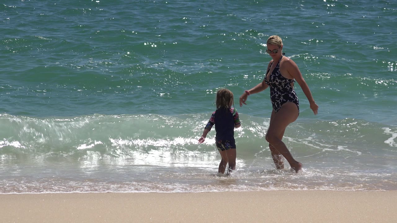 The woman steps through shallow water beside the child while a low wave nears the sand in Cabo San Lucas, Mexico.