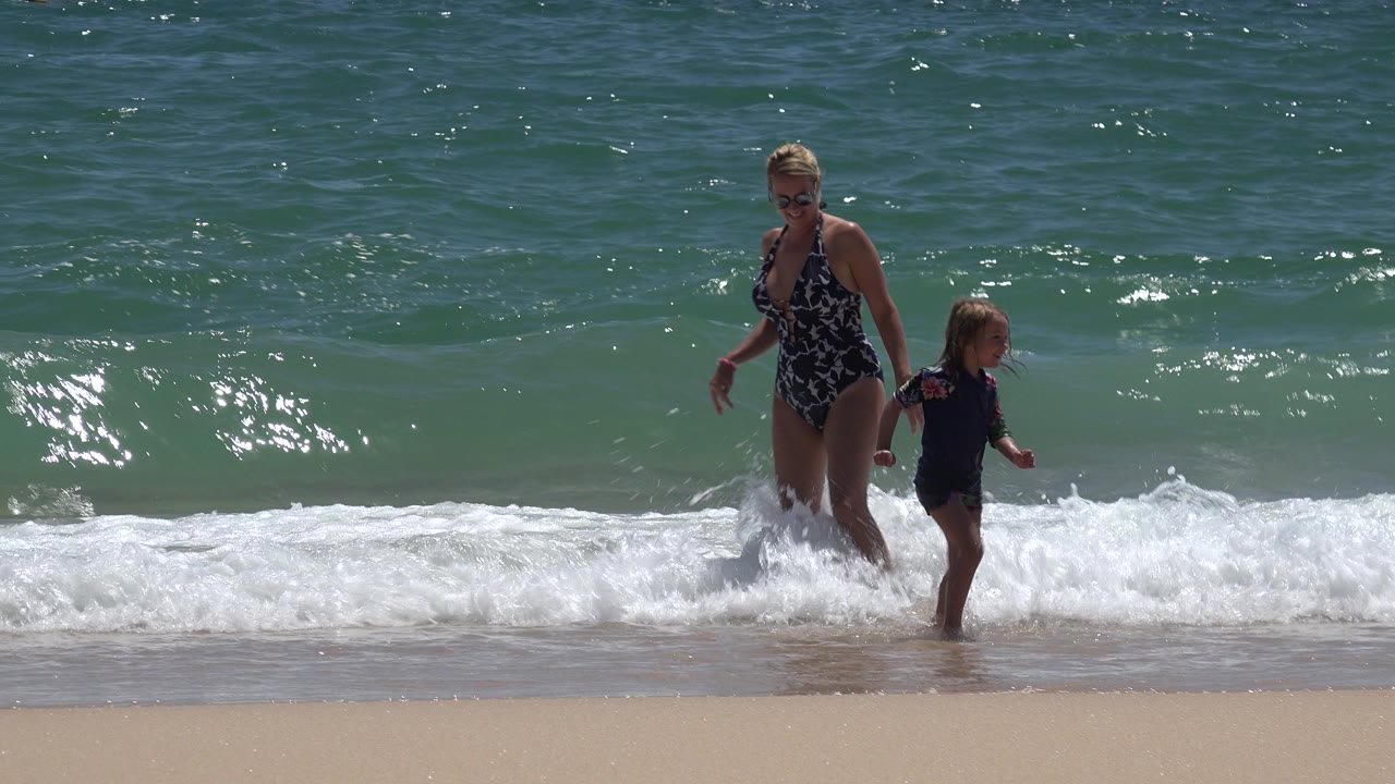 Woman and young child standing in shallow surf on a sunny beach.