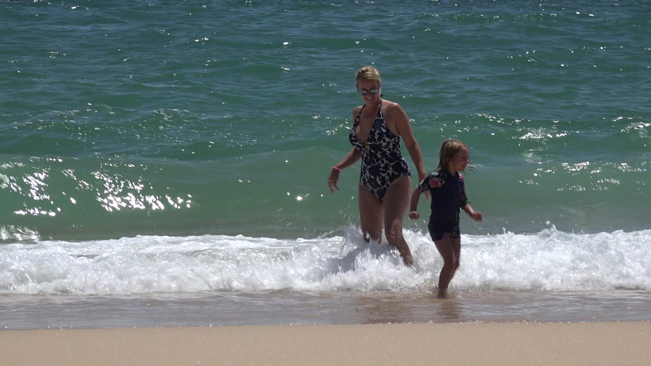 In Cabo San Lucas, Mexico, the woman and child move along the shoreline while a low wave breaks across the sand behind them.