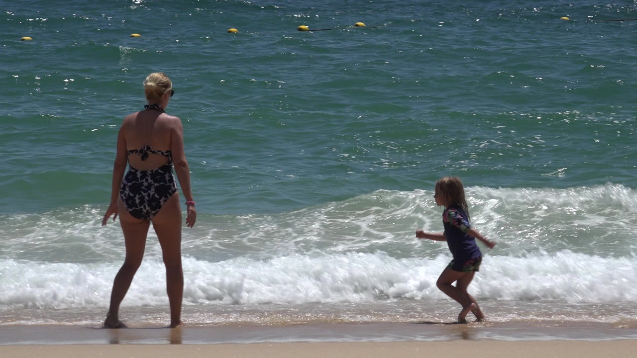 The woman faces the water as the child runs or steps along the foamy shoreline in Cabo San Lucas, Mexico, with the ocean surface bright under direct sun.