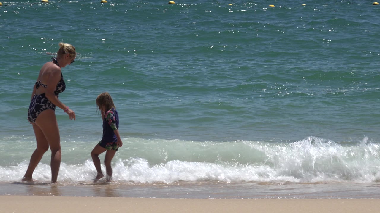 In Cabo San Lucas, Mexico, the woman faces the child as a foamy wave breaks between them on the sandy shoreline.