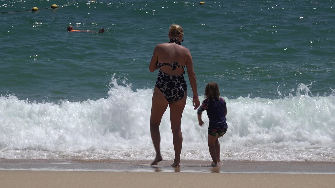 Woman and child stand at the edge of breaking waves.