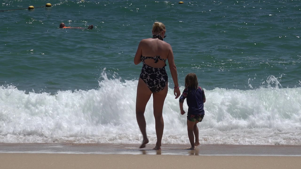 In Cabo San Lucas, Mexico, the woman and child remain near the waterline as white foam spreads across the sand and small swimmers are visible offshore.