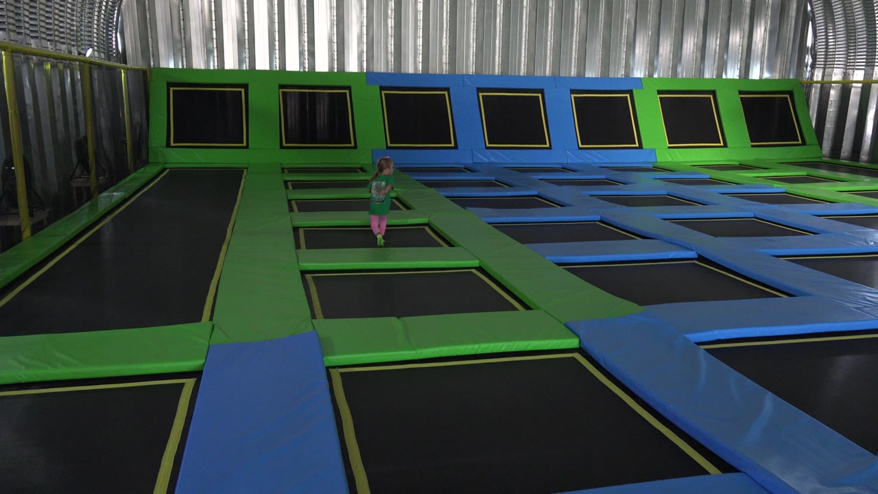 At 0:01, the child remains in the midground on a green walkway, framed by black trampoline rectangles in the foreground and a row of angled wall trampolines behind her.