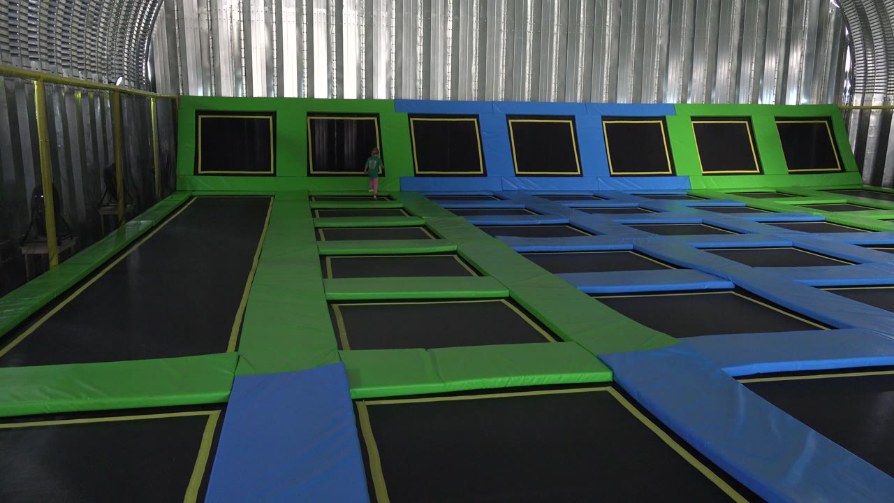 A small child becomes visible near the back trampoline wall in the Cabo San Lucas indoor play space, framed by green pads, blue pads, and black trampoline panels.