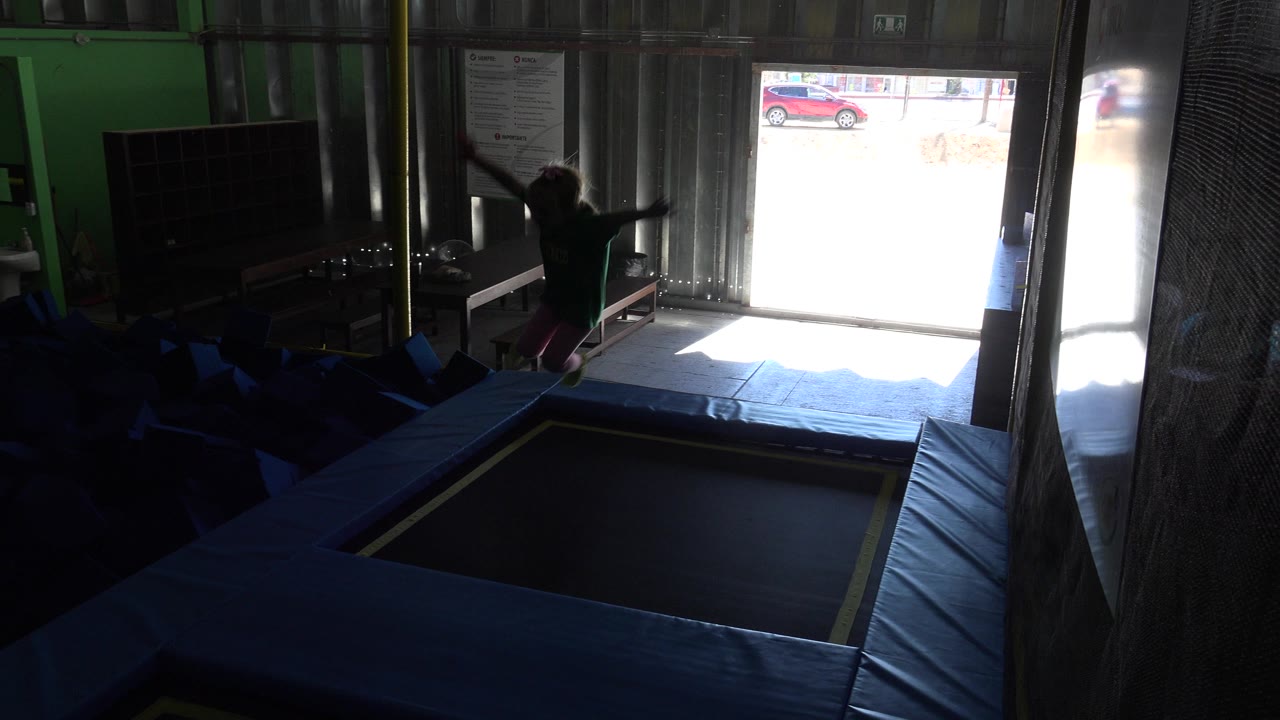 The child remains near the trampoline edge in Cabo San Lucas, Mexico, framed by blue pads, a dark play area, and a blown-out doorway showing a red car outside.