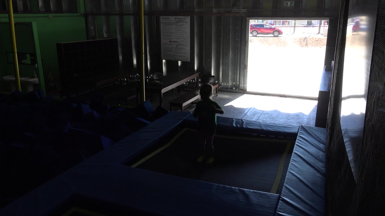A child stands near blue padded trampoline mats in a dim indoor play area in Cabo San Lucas, Mexico, with bright daylight spilling through an open doorway.