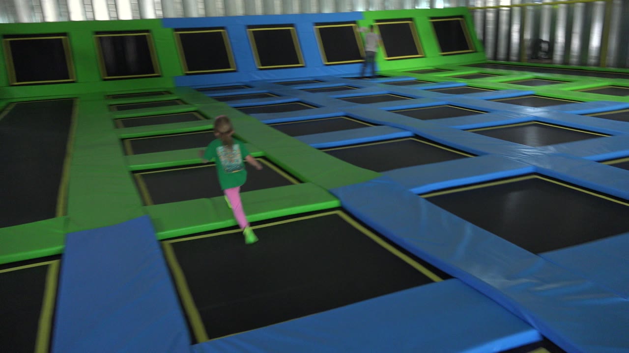 The girl remains near the foreground trampoline lane while rows of black jumping surfaces and angled wall trampolines fill the Cabo San Lucas indoor play space.