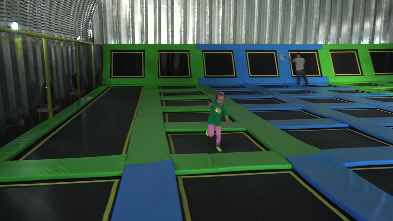 Young girl moves forward on a green lane in the Cabo San Lucas trampoline park, with repeated blue and black trampoline sections and a distant adult near the back wall.