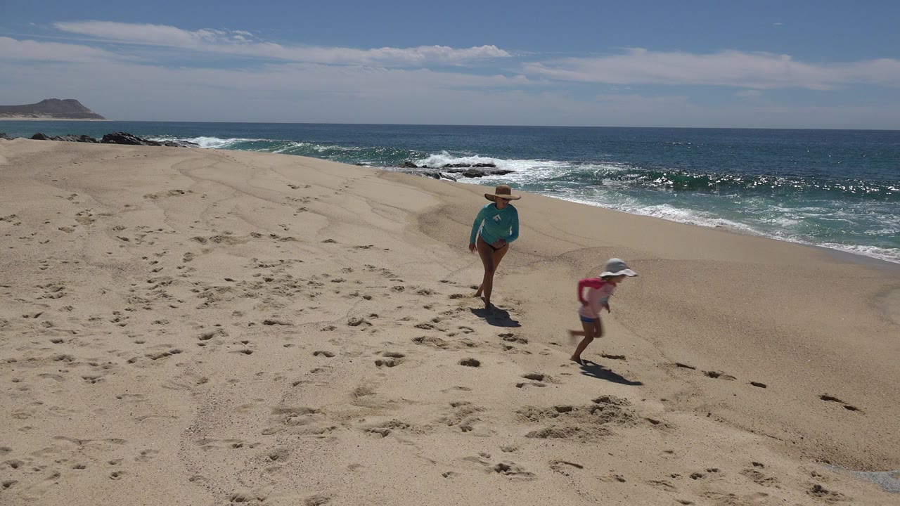 At 0:00 in Cabo San Lucas, Mexico, a mother in a turquoise top and straw hat stands on open sand while her young daughter in a pink top and white hat runs near the surf.