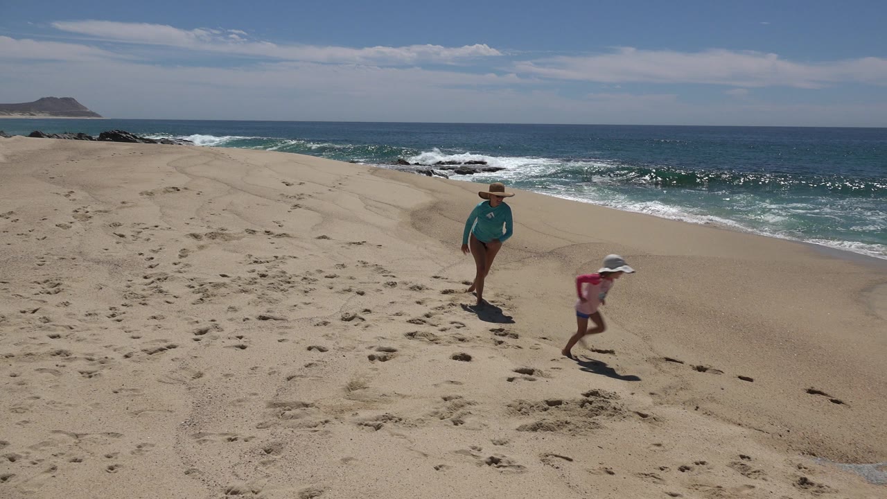 At 0:01 in Cabo San Lucas, Mexico, the child runs ahead across footprint-marked sand as the mother follows, with waves breaking along the beach in the background.