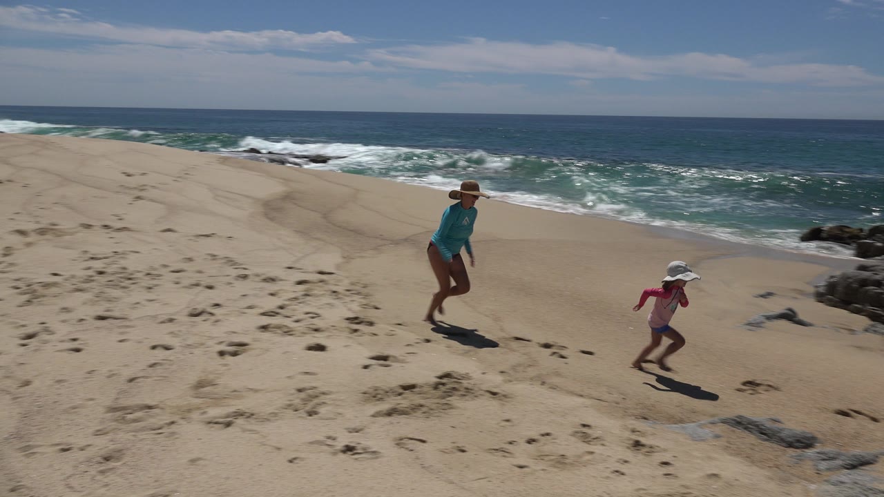 Mother and child run along a beach with surf and rocks nearby.