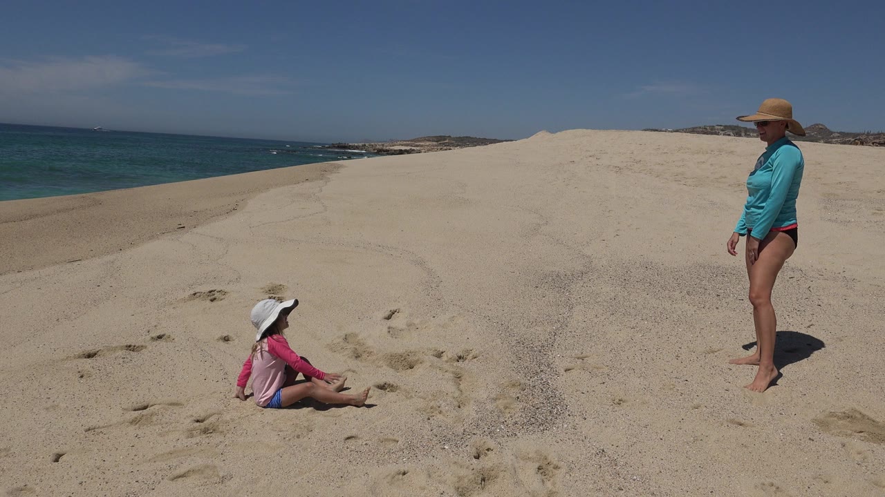 At 0:01 in Cabo San Lucas, the woman remains standing on the dune while the child sits farther left in the sand, framed by bright beach light, ocean water, and low coastline.