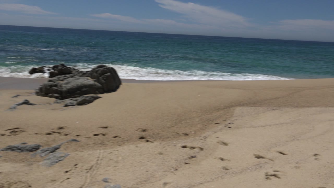Cabo San Lucas beach view with a large dark rock cluster left of center, pale sand in front, breaking surf behind it, and no visible people.