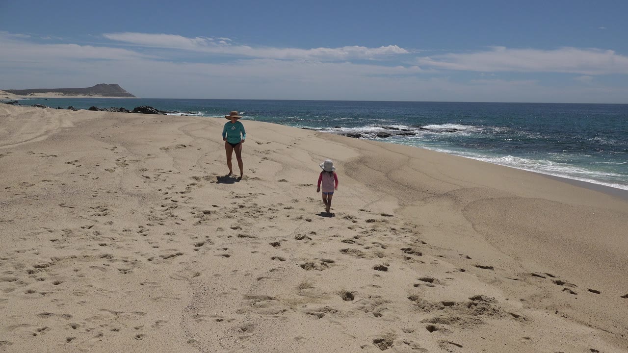 In Cabo San Lucas, Mexico, the mother remains near center while the child stands closer to the right, with white waves breaking along the beach in the background.