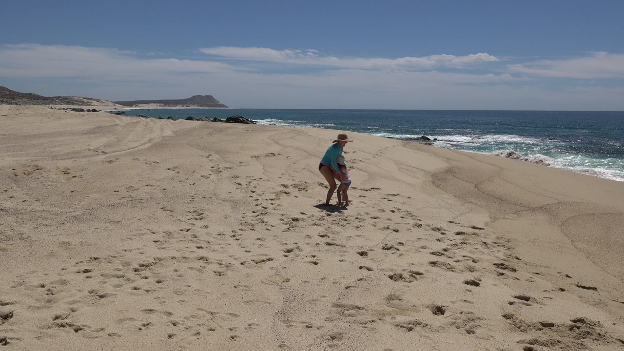 The woman and child remain near the center of the sandy Cabo San Lucas beach as the surf line and dark rocks sit behind them under bright sun.