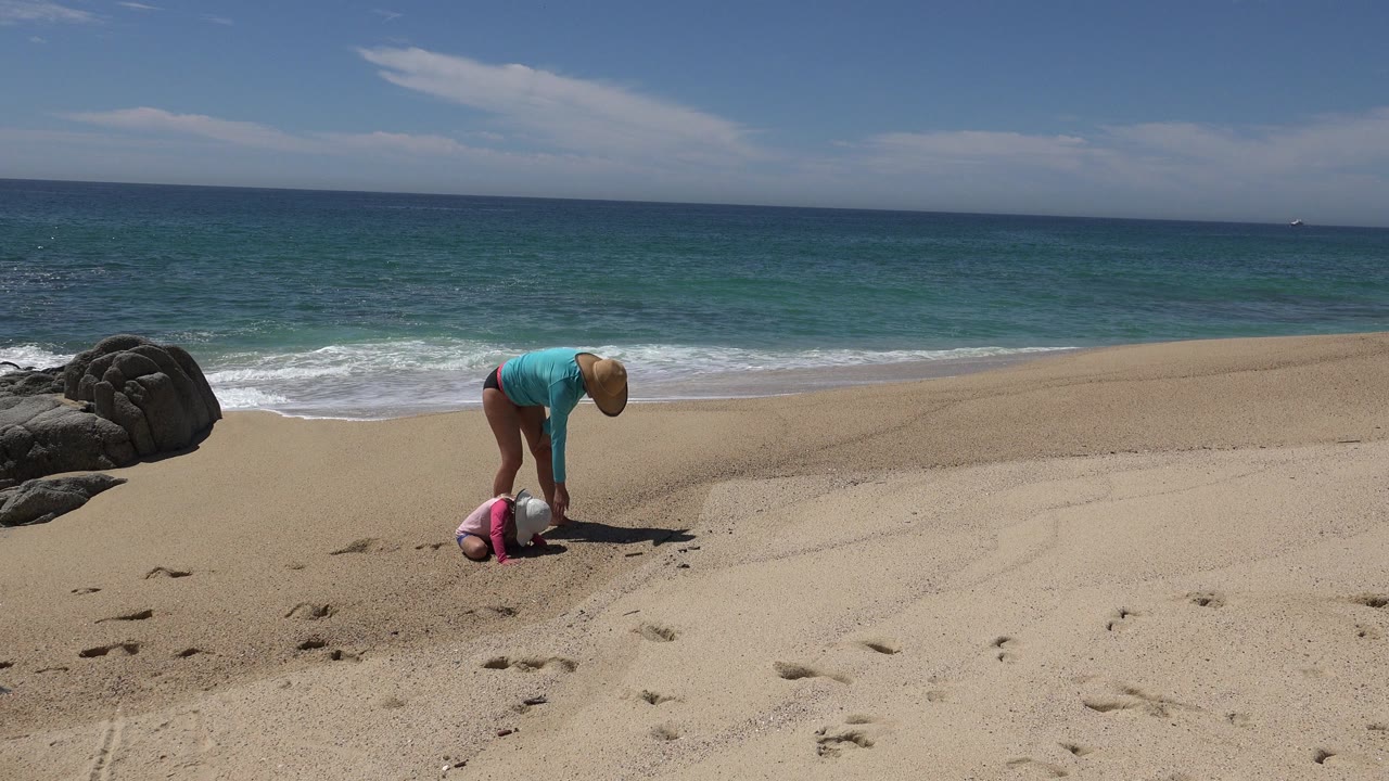 Adult bends over child on sandy beach near surf.