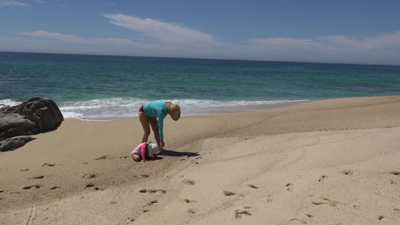 The adult remains bent toward the sand while the child stays low beside them on the Cabo San Lucas beach, with a breaking wave farther back.