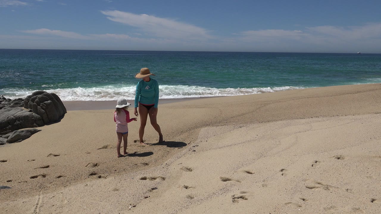 In Cabo San Lucas, Mexico, an adult in a wide hat and a small child in a white hat stand on pale sand near dark rocks with turquoise water and a blue sky behind them.