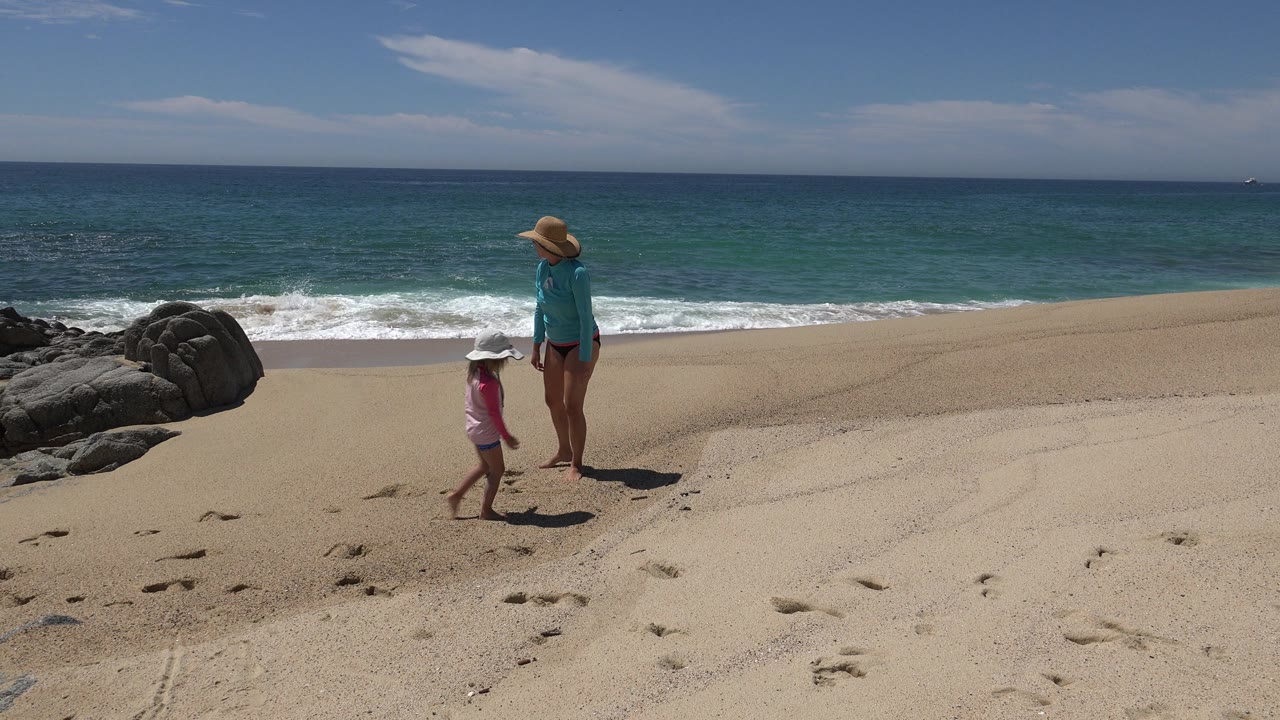 Adult and child walking across sand beside a rocky beach.
