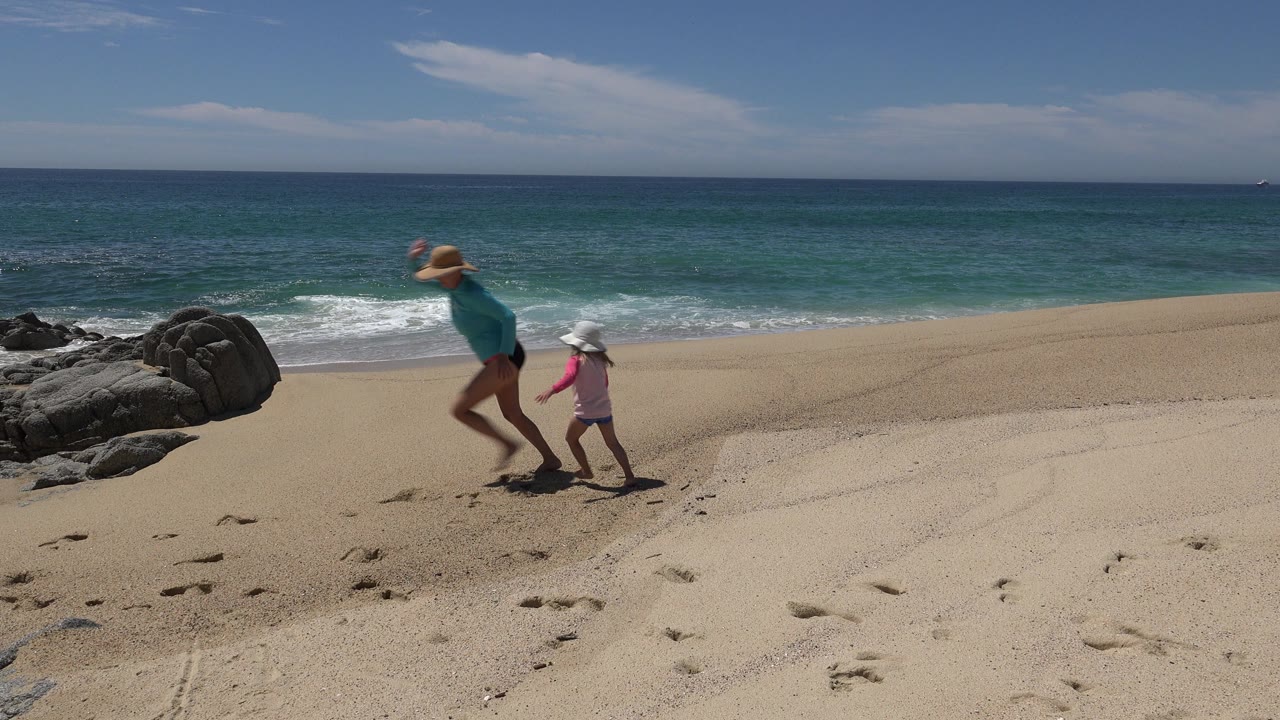 The mother and young girl continue running hand in hand on the Cabo San Lucas sand, with the rock outcrop closer behind them and the ocean horizon level in the background.