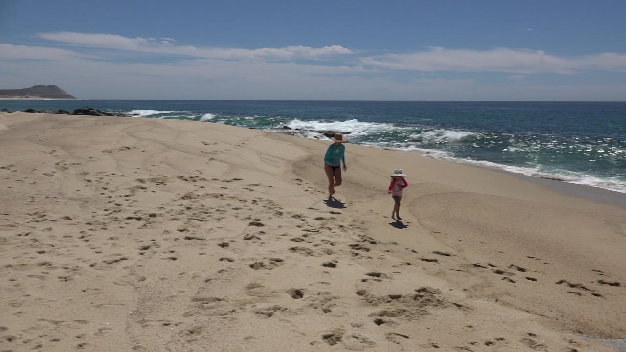 The mother and child move farther along the Cabo San Lucas shoreline, still framed wide against tan sand, white surf, blue water, and a low rocky landform in the distance.