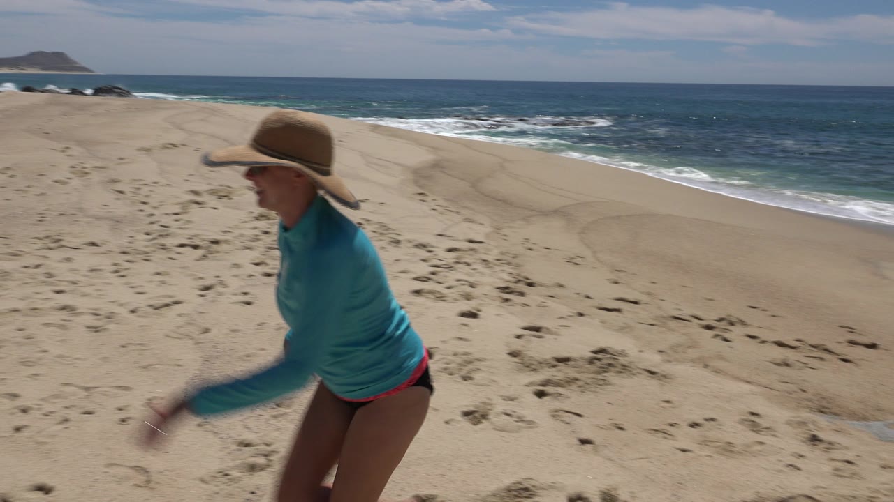 The woman continues across the foreground on the Cabo San Lucas sand while white surf lines the blue water behind her.