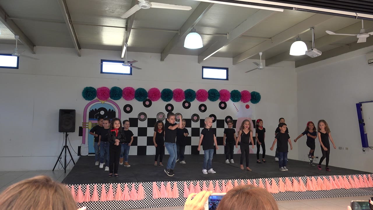 At 0:00, children in black shirts stand and dance across a decorated indoor stage in Cabo San Lucas, Mexico, with a checker backdrop, orange fringe, and audience heads at the bottom of frame.