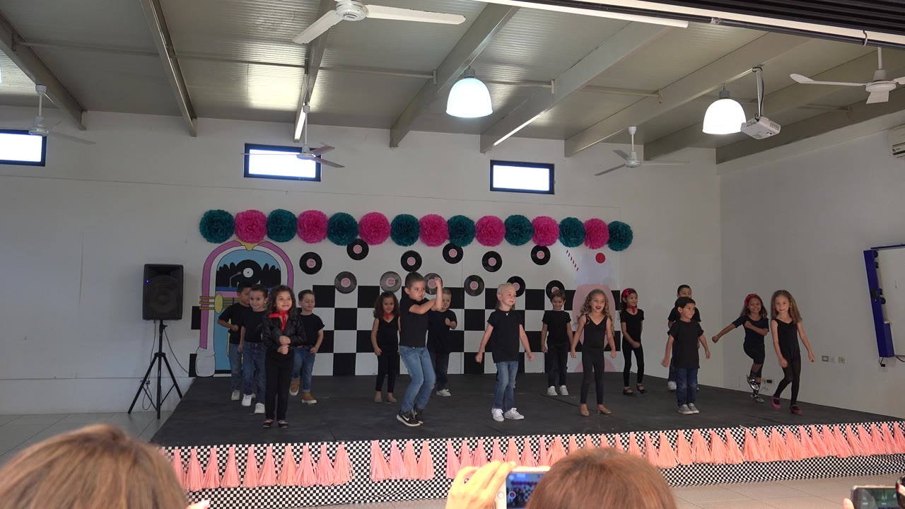At 0:01, the children shift positions on the Cabo San Lucas indoor stage while a boy near center steps forward and other dancers hold places in front of the checker backdrop.