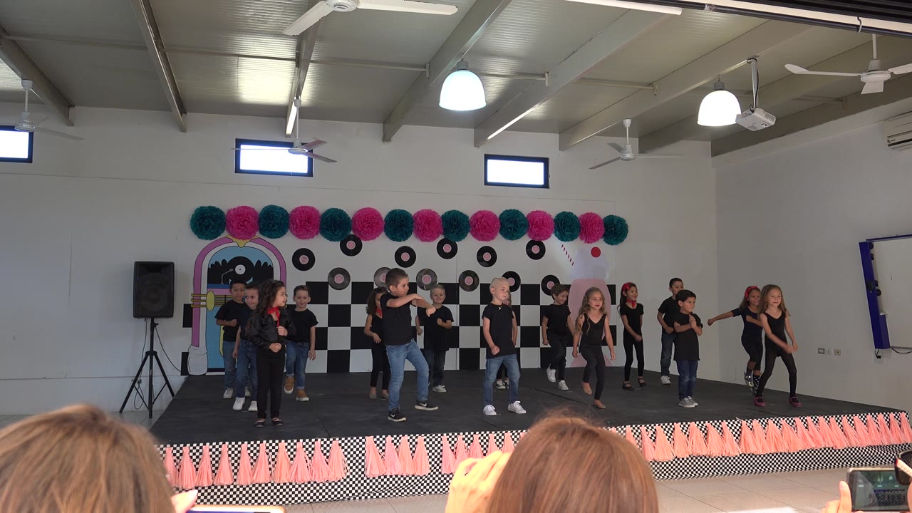 Kids perform on an orange-trimmed stage as audience members record.