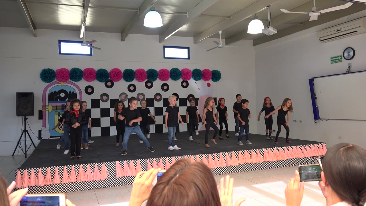 Children dancing on a decorated indoor stage while adults film from the audience.