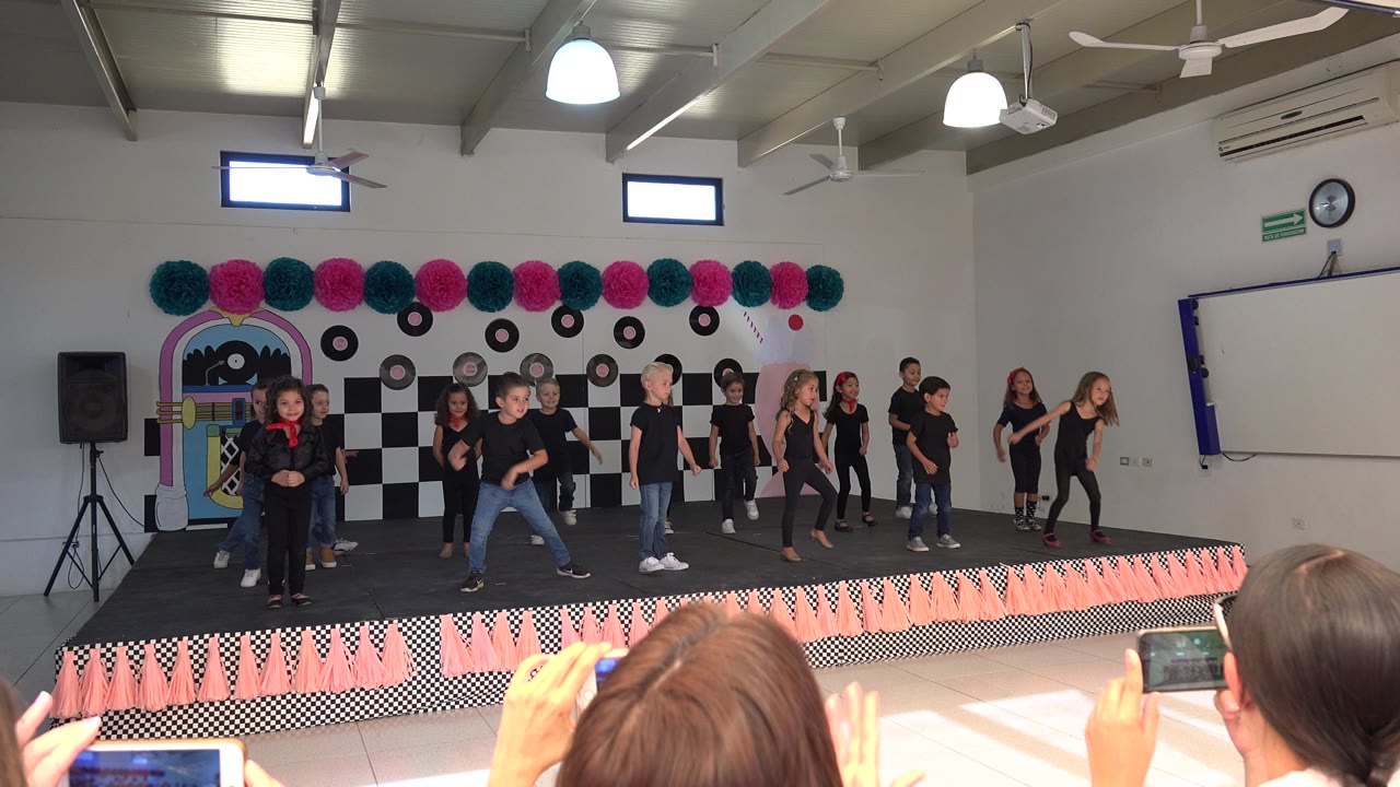 The group of children spreads across the Cabo San Lucas stage, with several dancers stepping forward and adults holding phones in the foreground.