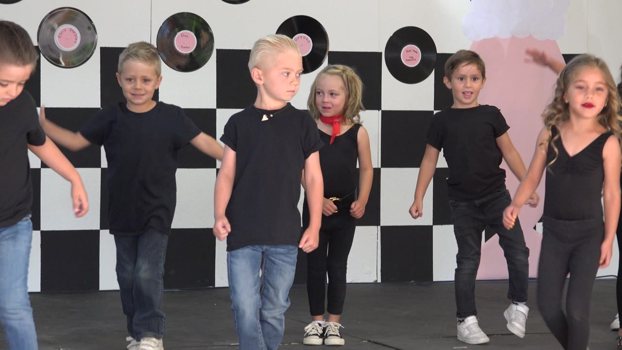 Children in black shirts and jeans move across a Cabo San Lucas stage with a checkerboard backdrop, pink decor, and record decorations behind them.