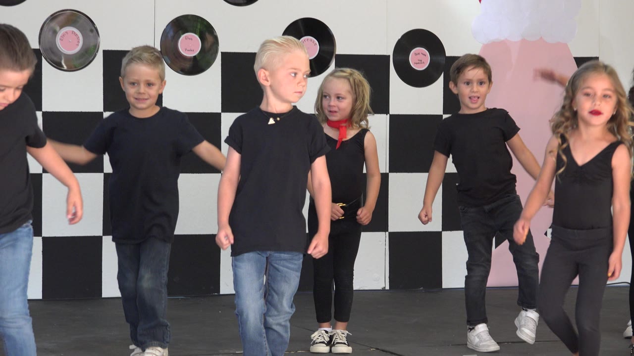 A blond boy, a girl with a red neck scarf, and other children stand mid-routine on the Cabo San Lucas stage in front of black-and-white panels.