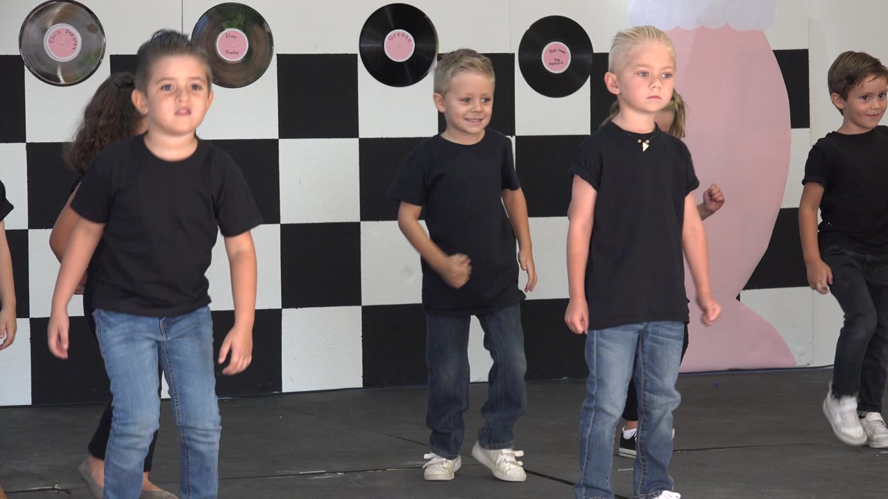 A young girl stands near the front while boys and girls behind her continue the Cabo San Lucas stage routine against record decorations and a checkerboard wall.