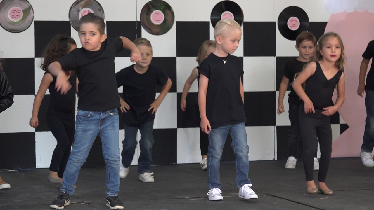 Young children in black tops and jeans practice dance steps on a stage in Cabo San Lucas, Mexico, with record decorations above a checkerboard backdrop.