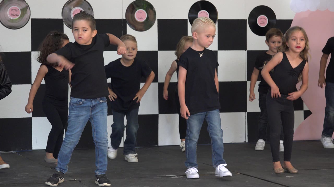 A boy in a black shirt and jeans stands near the front while other children move behind him in the Cabo San Lucas dance studio setting.